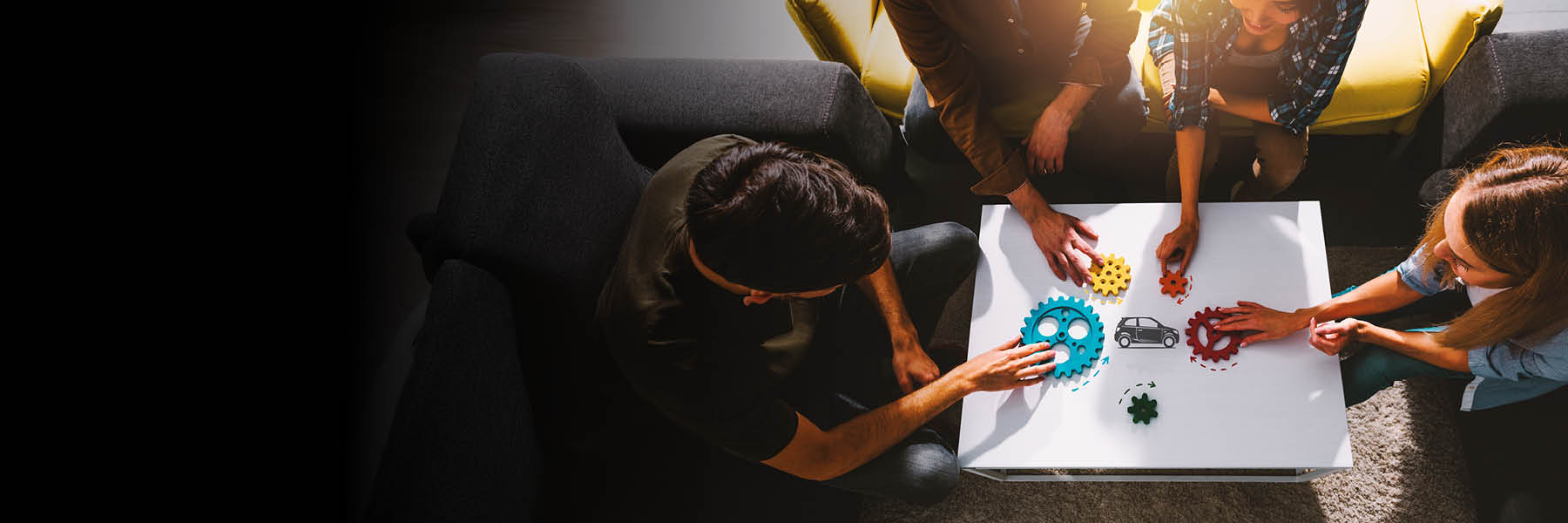 Mehrere Personen sitzen um einen Tisch und arbeiten gemeinsam an verschiedenfarbigen Zahnrädern, die auf einer Grafik mit einem Auto platziert sind, was auf Teamarbeit oder technische Planung hindeutet.