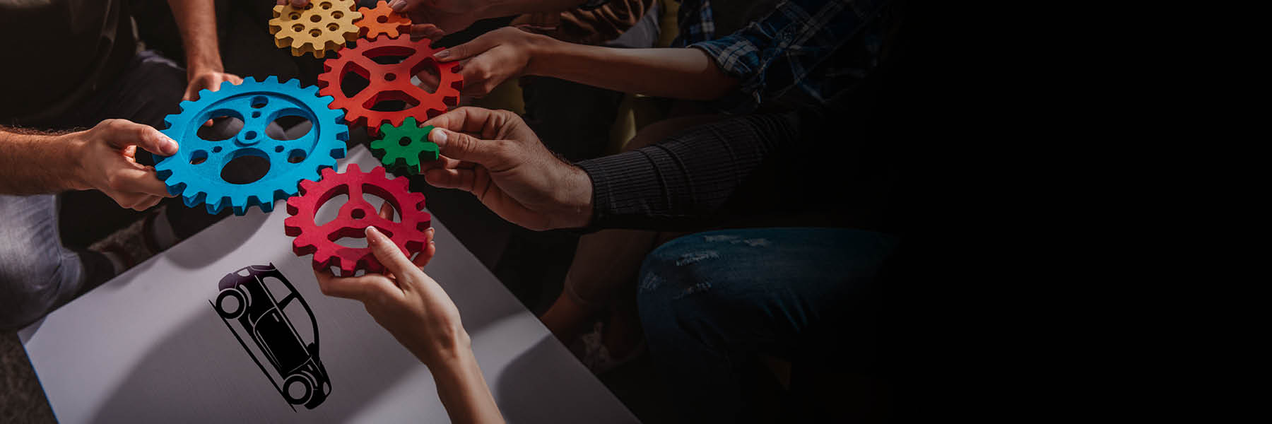 Mehrere Menschen sitzen zusammen und halten bunte Zahnräder in den Händen, die sie ineinandergreifen lassen – ein Symbol für Teamarbeit und Zusammenarbeit.