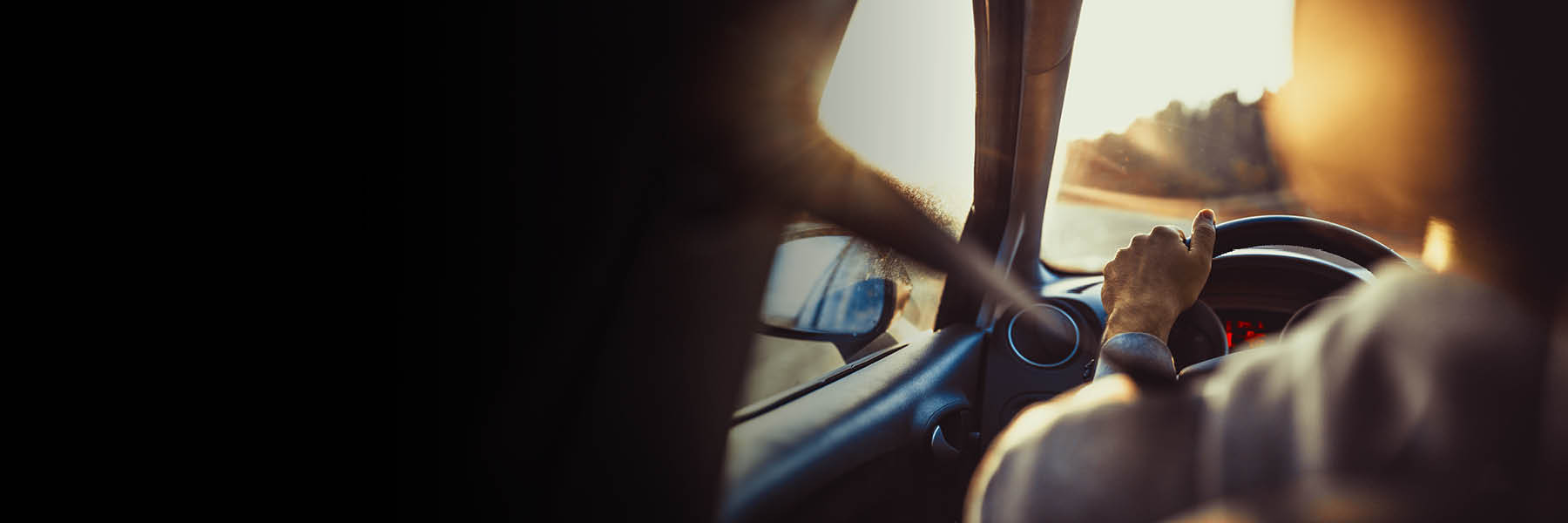 Eine Person fährt bei Sonnenuntergang ein Auto und hält das Lenkrad mit einer Hand, während die andere Hand am Fenster ruht.